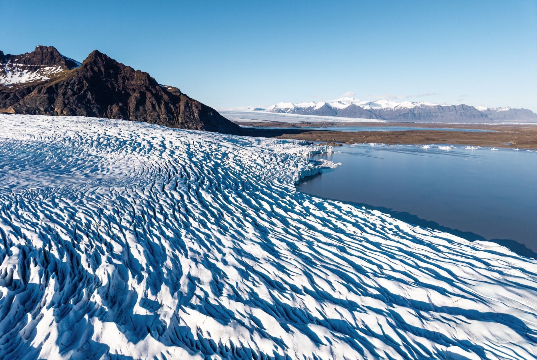 High-angle view of the textured, crevasse-filled ice of Vatnajökull Glacier alongside a glacial lake and rugged mountains.