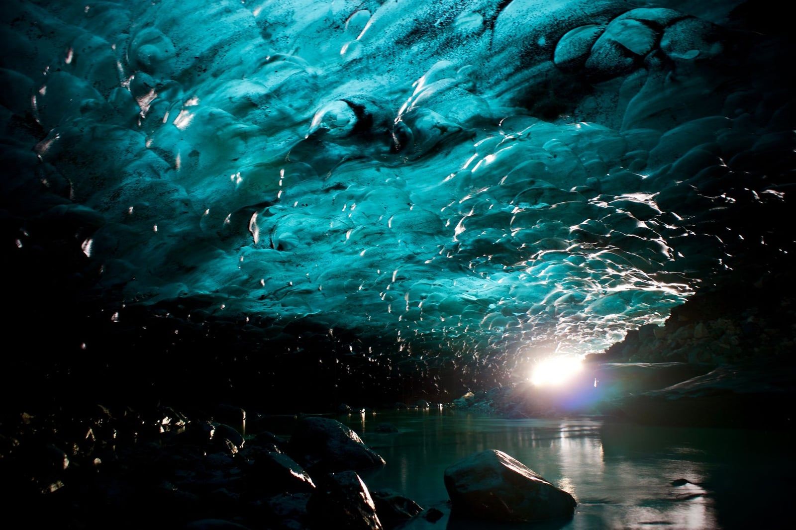 Sunlight streams into a natural blue ice cave with a scalloped frozen ceiling and reflective water pool at Vatnajökull Glacier in Iceland.